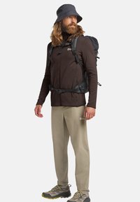 Homme portant une veste zippée marron foncé, un pantalon beige et des chaussures de randonnée. Il a un sac à dos noir et un chapeau seau noir.