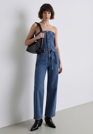 Donna che indossa una tuta di jeans senza spalline con bottoni sul davanti e cintura, stivaletti neri e che porta una borsa a tracolla nera davanti a un muro semplice.