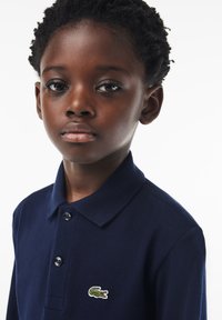 Tummansininen polo-paita, joka on valmistettu tekstuuri kankaasta, jossa on kolme nappia edessä ja pieni brodeerattu krokotiililogo rinnassa.