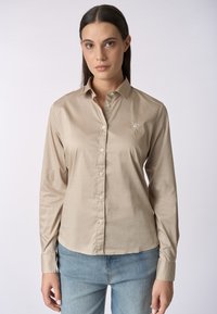 Camicia a maniche lunghe color beige chiaro realizzata in tessuto liscio, con chiusura frontale a bottoni e un piccolo dettaglio ricamato sul petto. Abbina a jeans blu.