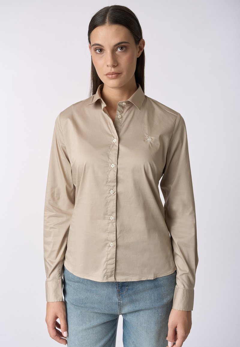 Camicia a maniche lunghe color beige chiaro realizzata in tessuto liscio, con chiusura frontale a bottoni e un piccolo dettaglio ricamato sul petto. Abbina a jeans blu.