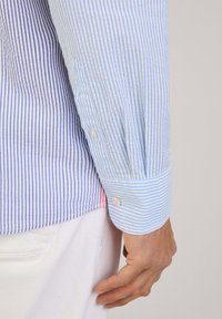 Camisa a rayas azul y blanca con puños de botón. Tela de algodón, con rayas verticales, y un acento rosa en contraste cerca del dobladillo.