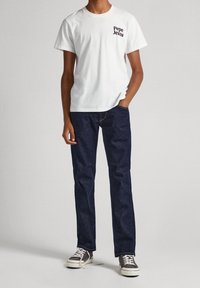 Camiseta de algodón blanca con "Pepe Jeans" en texto oscuro, combinada con jeans de mezclilla azul oscuro y zapatillas grises con suelas y cordones blancos.
