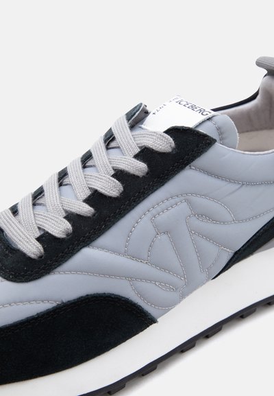 Γκρι και μαύρο αθλητικό παπούτσι με λευκά κορδόνια, μαύρες σουέντ λεπτομέρειες, κεντημένο λογότυπο και λευκή σόλα σε φωτεινό φόντο.