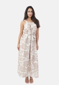 Kvinde står iført en hvid ærmeløs maxikjole med beige blomster mønstre og et taljebånd, kombineret med sandaler med klare stropper.