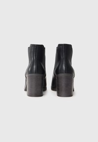Botas de cuero negro hasta el tobillo con un acabado suave, punta redondeada y un tacón grueso gris. Las costuras verticales en la parte trasera añaden detalle.