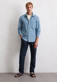 Chemise bleu clair à manches boutonnées, pantalon marine et sandales noires. La chemise a une poche et des manches roulées ; le pantalon est slim-fit, créant un look décontracté.