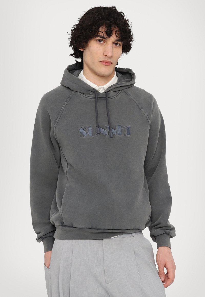 Sweat à capuche gris avec cordon de serrage et poignets côtelés, portant "SUNNEI" brodé en bleu sur le devant. Tissu doux et texturé.