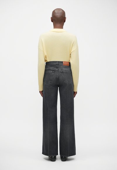 Un pull surdimensionné jaune clair et un jean taille haute gris foncé à jambes larges, avec une patch en cuir sur la ceinture arrière.