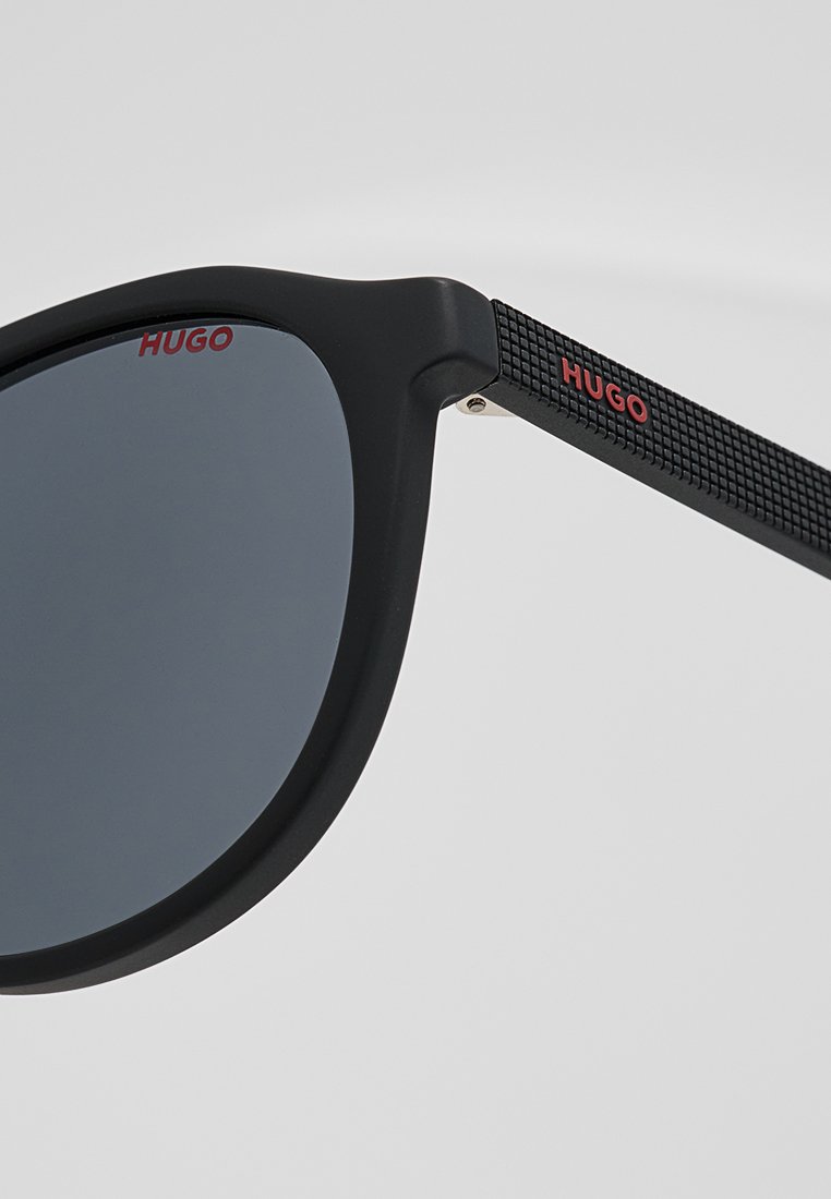 Schwarze Sonnenbrille mit mattem Finish; rechteckiger Rahmen und dunkle Gläser. Das Bügelstück zeigt ein strukturiertes Muster mit dem Markenlogo in Rot.