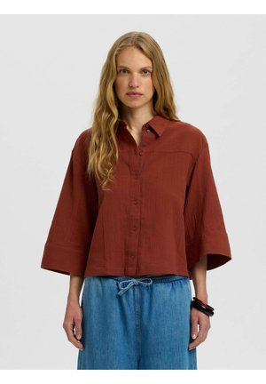 Femme aux longs cheveux ondulés porte une chemise ample couleur rouille avec boutons, manches larges et un pantalon en jean bleu clair à cordon.