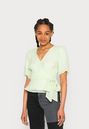 Blusa envolvente verde pálido com mangas curtas em balão, tecido texturizado, cintura amarrada e decote em V, combinada com calças pretas e cinzas.