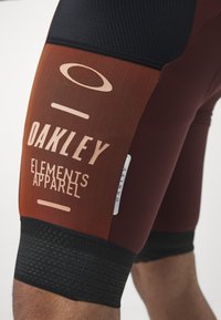 Gros plan sur la cuisse d'une personne portant un short de cyclisme Oakley marron et noir avec l'inscription "Oakley Elements Apparel" imprimée sur le tissu.