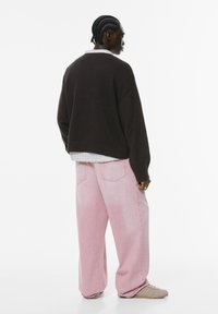 Brun oversized strikket sweater, lagd ovenpå en ternet skjorte, parret med lyserøde brede bukser. Fodtøjet er beige sneakers.