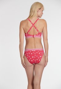 Rdež bikini komplet z belimi pikami, ki ima križno zasnovo in črtaste detajle. Tkanina se zdi gladka in raztegljiva.