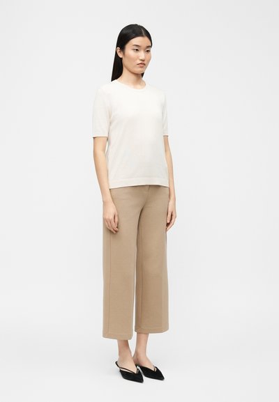 Γυναίκα που στέκεται με μακριά μαύρα μαλλιά, φοράει λευκό τοπ με κοντά μανίκια, μπεζ παντελόνια φαρδιά και κοντά, και μαύρα μυτερά παπούτσια με λουράκι στην φτέρνα.