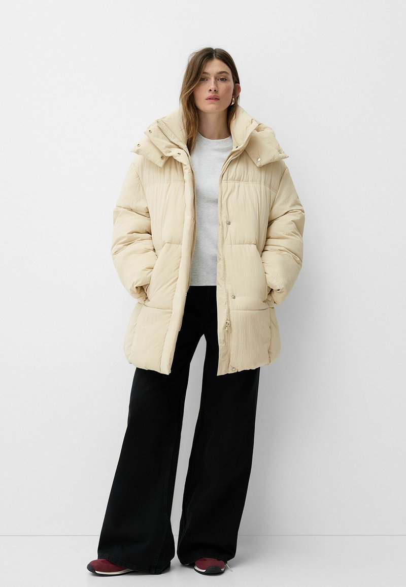 Donna con lunghi capelli castani, che indossa un giubbotto beige imbottito, una camicia bianca, pantaloni larghi neri e sneakers rosse, in piedi contro uno sfondo bianco.