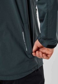Gros plan d'une main partiellement dans la poche latérale d'une veste noire étiquetée "Swedemount On Course" avec un poignet réglable visible.
