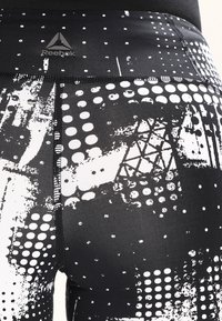 Legíny Reebok s černým základem a bílými abstraktními vzory, včetně teček a geometrických tvarů, vyrobené z hladkého a elastického materiálu.