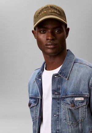 Hellblaue Jeansjacke mit zwei Brusttaschen, getragen über einem weißen T-Shirt. Olivgrüne Mütze mit eingesticktem Text "Calvin Klein New York."