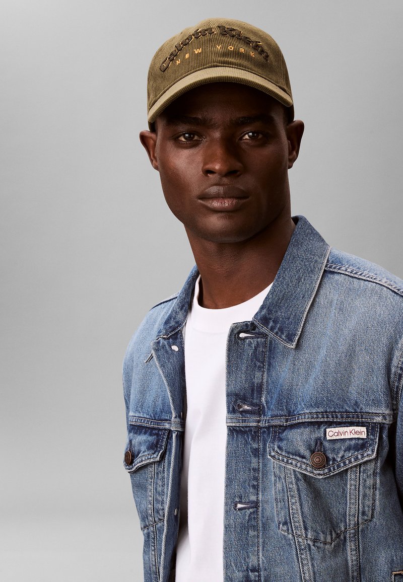 Veste en denim bleu clair avec deux poches poitrine, portée sur un t-shirt blanc. Casquette vert olive avec texte brodé "Calvin Klein New York".