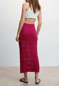 Gehäkelter Maxirock in kräftigem Pink mit offenem, gitterartigem Muster. Kombiniert mit einem weißen Crop-Top. Schwarze Sandalen runden das Outfit ab.