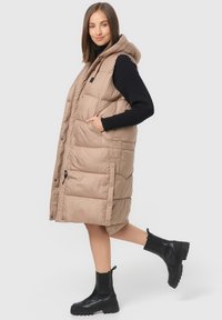 Gilet matelassé beige avec une capuche, doté de boutons-pression, de poches latérales et d'un design matelassé. Porté par-dessus un pull noir côtelé et des bottes noires.