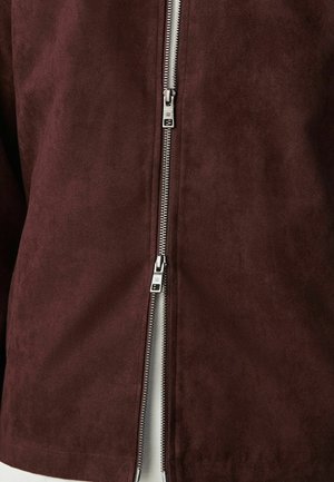 Doppelter silberner Reißverschluss, teilweise geöffnet, an einer dunkelbraunen Wildlederjacke, die den darunterliegenden hellen Stoff zeigt.