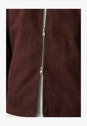 Double silver zipper partially unzipped on dark brown suede jacket, showing light fabric underneath.