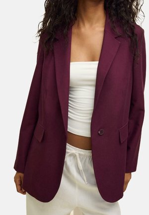 Frau trägt einen dunkelvioletten Blazer über einem weißen Bandeau-Top und weißen Hosen mit Kordelzug, gezeigt vom Hals bis zu den Oberschenkeln.