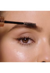 Applicateur de gel pour les sourcils avec une brosse à poils noirs en utilisation. Gros plan sur un œil marron et une peau lisse avec une légère illumination. Cils marron visibles.