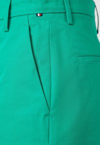 Grüne maßgeschneiderte Hosen aus glattem, strukturiertem Stoff, mit Falten und einer Gesäßtasche. Kleines farbblockiertes Etikett am Bund.