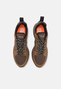 Brown athletic shoes with mesh and suede textures. Features black and white laces, orange insole, and reflective accents on the sides.