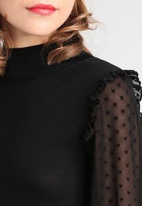 Schwarzes Strickoberteil mit hohem Kragen, das durchsichtige Ärmel mit kleinen schwarzen Herzmustern und Rüschensäumen aufweist.