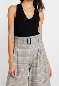 Czarny bezrękawnik z głębokim dekoltem w kształcie litery V. Spodnie w kratę z wysokim stanem w beżowe, niebieskie i brązowe kolory, z szerokim paskiem z prostokątną klamrą.