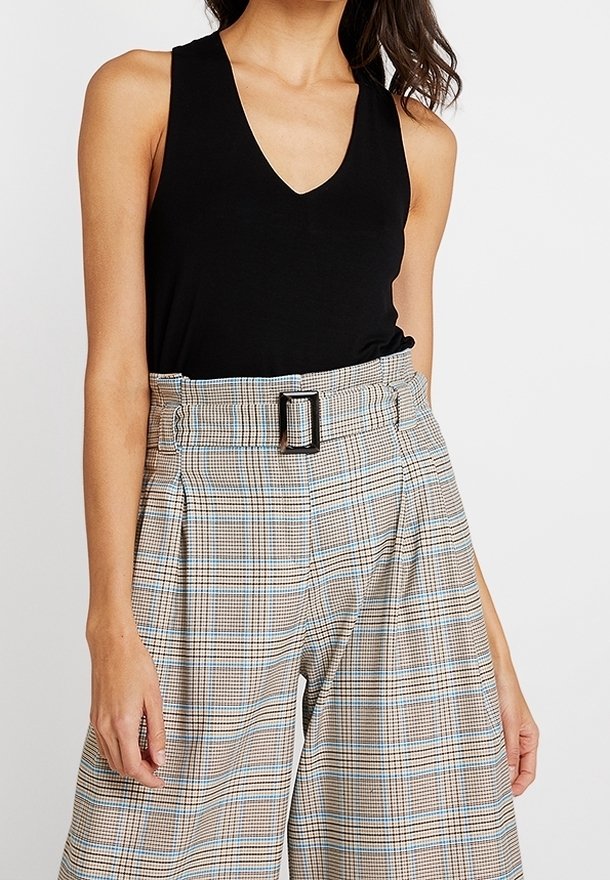 Czarny bezrękawnik z głębokim dekoltem w kształcie litery V. Spodnie w kratę z wysokim stanem w beżowe, niebieskie i brązowe kolory, z szerokim paskiem z prostokątną klamrą.