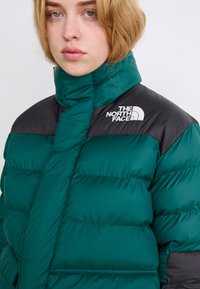 Mørkegrønn puffjakke med høy krage, svarte skulderdetaljer og hvit logo. Stoffet ser ut til å være quiltet og skinnende med synlige sømmer.