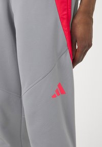 Detailní záběr na osobu v šedých sportovních kalhotách s červenými bočními akcenty a červeným logem u kolena, ruka položená na boku.