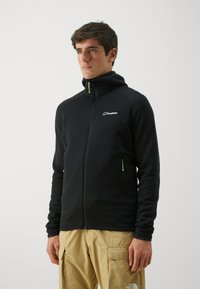 Mlad muškarac nosi crnu Berghaus duksericu sa patent zatvaračem i neon zelenim povlačenjem, uz bež cargo hlače iz The North Face, na jednostavnoj pozadini.