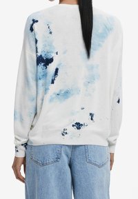 Pull blanc avec des motifs abstraits bleu clair et bleu marine, coupe décontractée, poignets et ourlet côtelés. Associé à un pantalon en jean bleu clair.