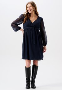 Navyblaues Kleid mit tailliertem Oberteil, tiefem V-Ausschnitt und langen, durchsichtigen Ärmeln. Ein passender gebundener Gürtel betont die Taille. Kombiniert mit knielangen schwarzen Stiefeln.