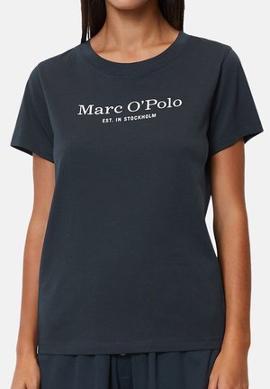 Eine Frau trägt ein dunkelblaues Kurzarm-T-Shirt mit der Aufschrift "Marc O'Polo EST. IN STOCKHOLM" in Weiß auf der Brust.