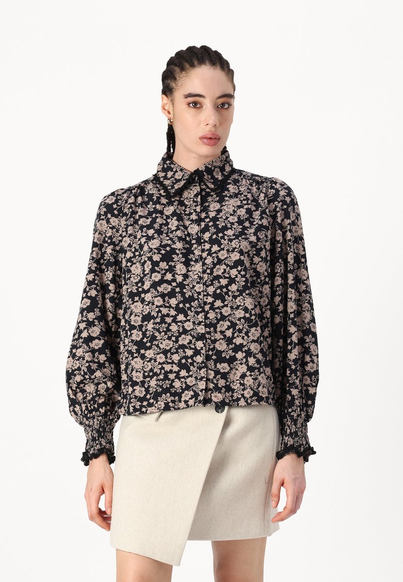 Blusa negra con flores en tonos rosa apagado, de mangas largas y abullonadas, y cuello con solapa. Combinada con una falda envolvente de un beige claro.