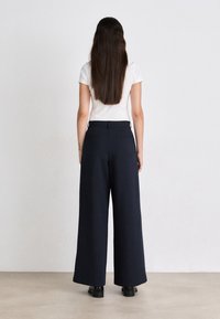 Pantaloni larghi blu scuro in tessuto, caratterizzati da una texture liscia, tasche posteriori e vita alta, abbinati a un top bianco aderente.