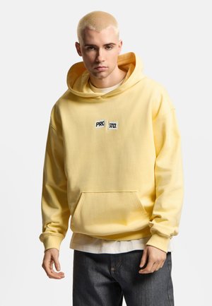Młody mężczyzna z krótkimi blond włosami, ubrany w żółtą bluzę z kapturem z logo "PROSTO." oraz ciemne dżinsy na jednolitym tle.