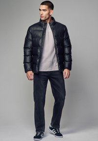 Chaqueta de plumas negra con un cuello alto, combinada con una sudadera gris claro, jeans oscuros y zapatillas negras, en un fondo neutro.