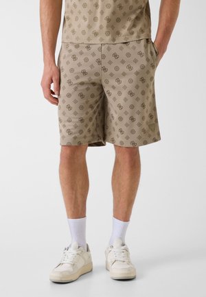Mann trägt beige Shorts mit durchgehendem dunkelbraunem geometrischem und Logo-Muster, kombiniert mit weißen Socken und weißen Turnschuhen.