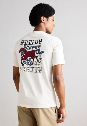 Mann som står med ryggen til iført hvit T-skjorte med en rød enhjørning og abstrakt blått og svart tekstdesign på baksiden, kombinert med beige bukser.