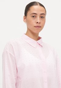 Pink button-up shirt with a collar, featuring a striped pattern and small sparkling accents throughout the fabric. Smooth texture, relaxed fit.