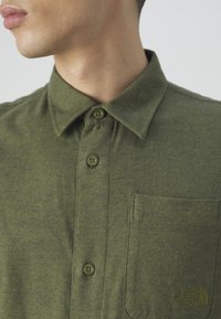 Camisa de franela verde con cuello, que presenta una tapeta de dos botones, bolsillo lateral y un pequeño logo bordado en el bolsillo. Textura suave.